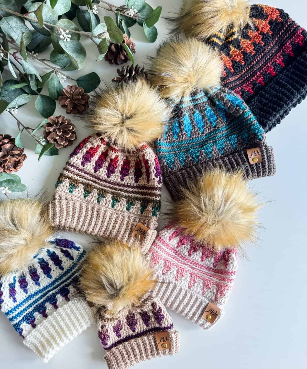 Seven knitted hats with pom-poms, featuring multicolored patterns, are displayed alongside eucalyptus branches and pinecones.