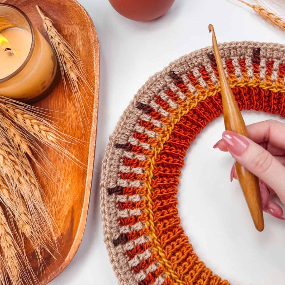 A person holds a wooden crochet hook above a circular crochet piece with orange and brown stripes. A lit candle and wheat stalks in a wooden dish are nearby.