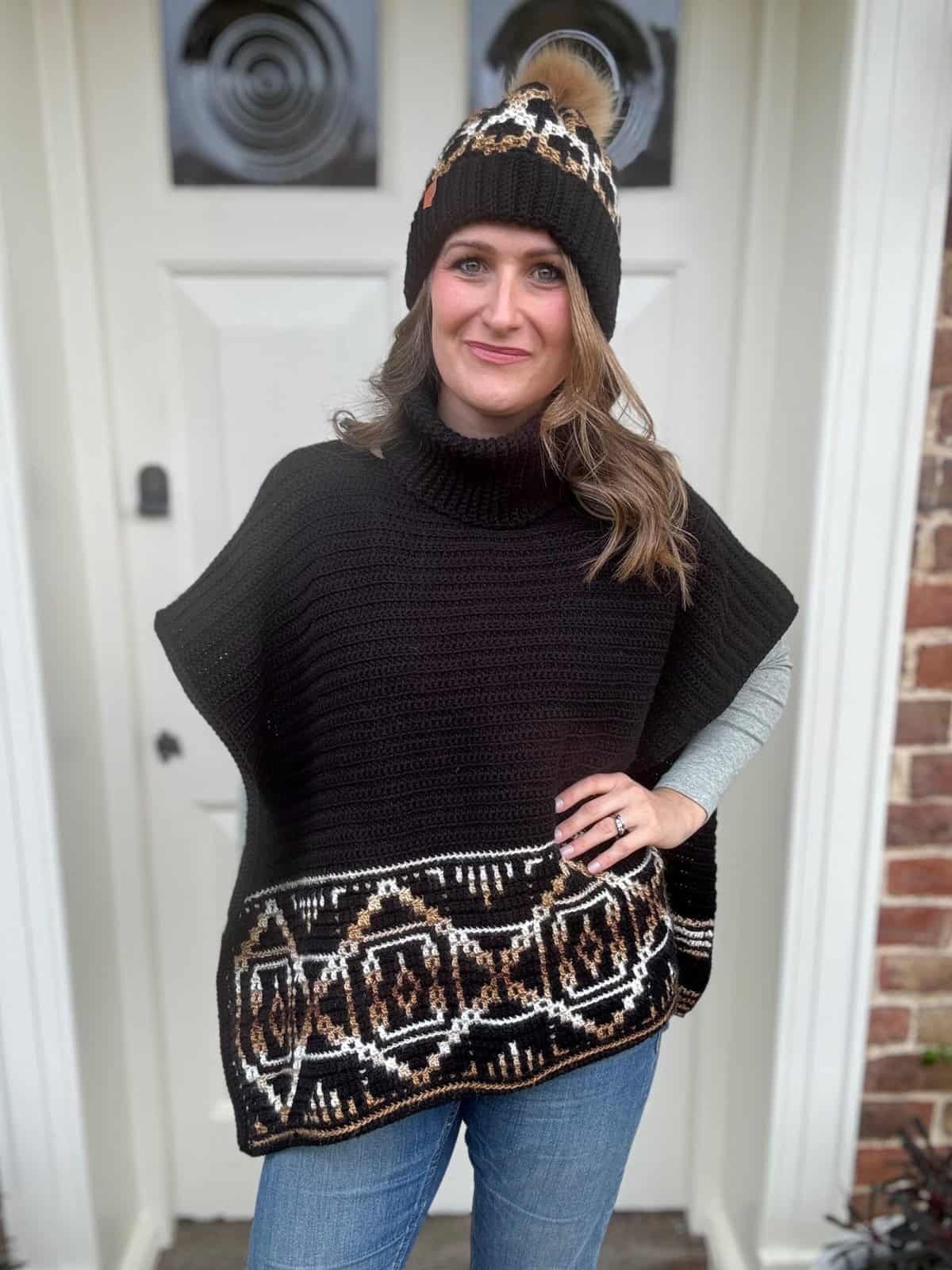 Person wearing a black knit poncho with geometric patterns and a matching beanie stands in front of a white door.