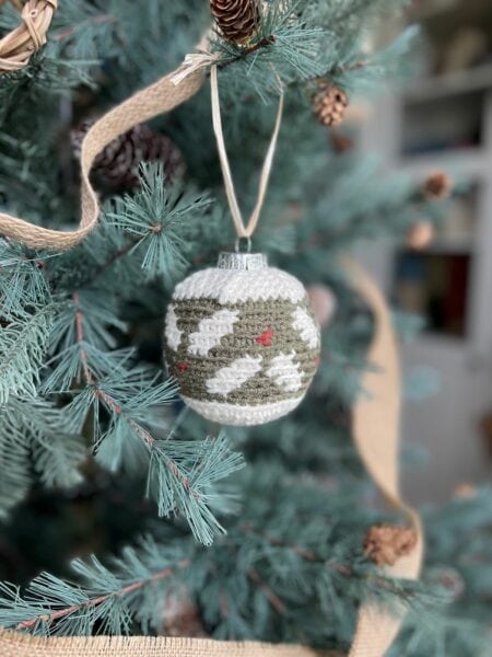 A green and white crochet bauble with bird and tree patterns hangs on a pine tree, adorned with burlap ribbon, pinecones, and hints of holly.