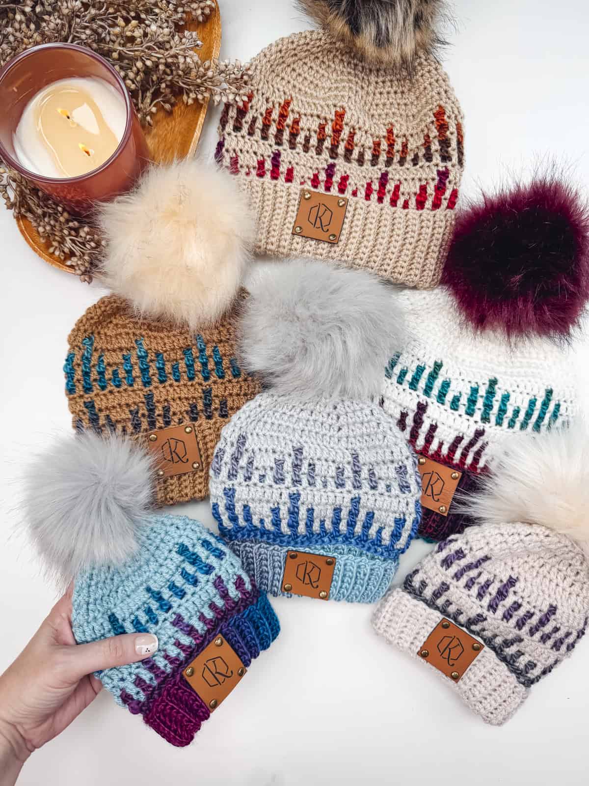 Seven knitted hats with pom-poms in various colors and patterns are arranged on a table next to a lit candle.