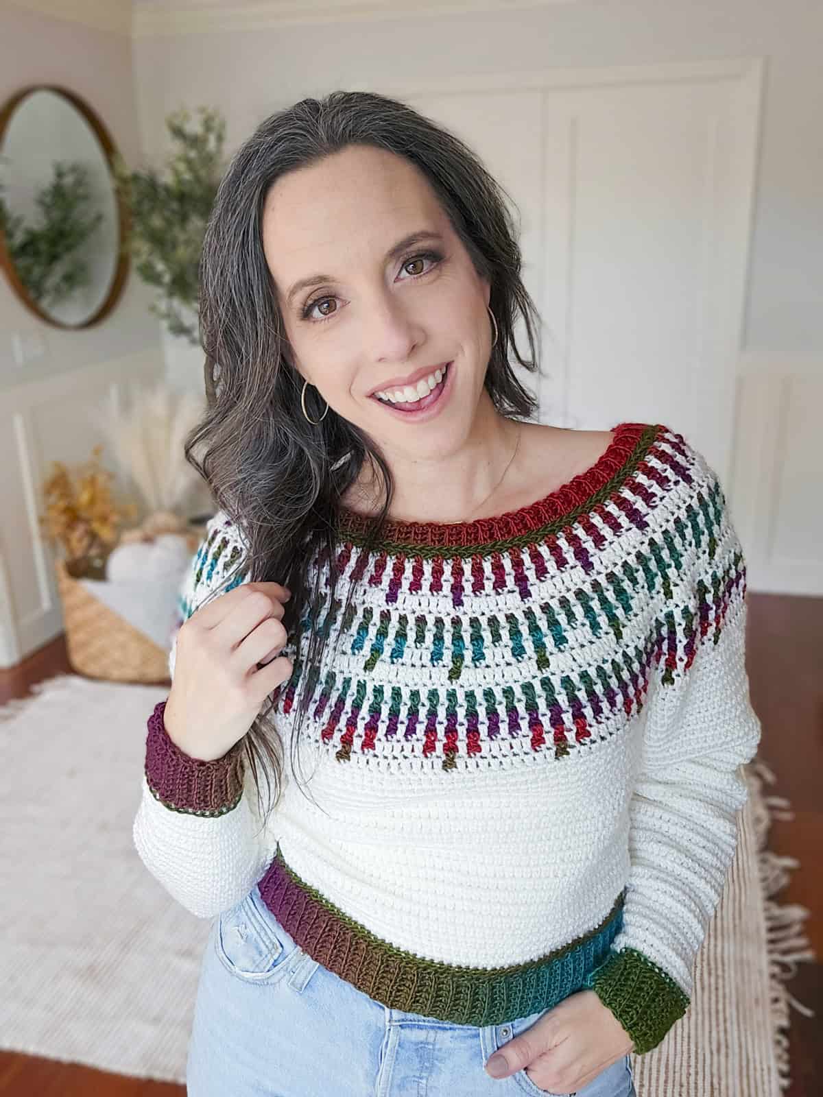 Person smiling indoors, wearing a knit sweater with colorful patterns. Light-colored room with a round mirror and a basket in the background.