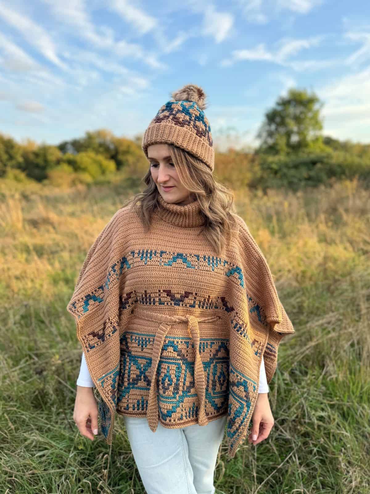 A person wearing a mosaic crochet hat and patterned poncho stands in a field with grass and trees under a blue sky.