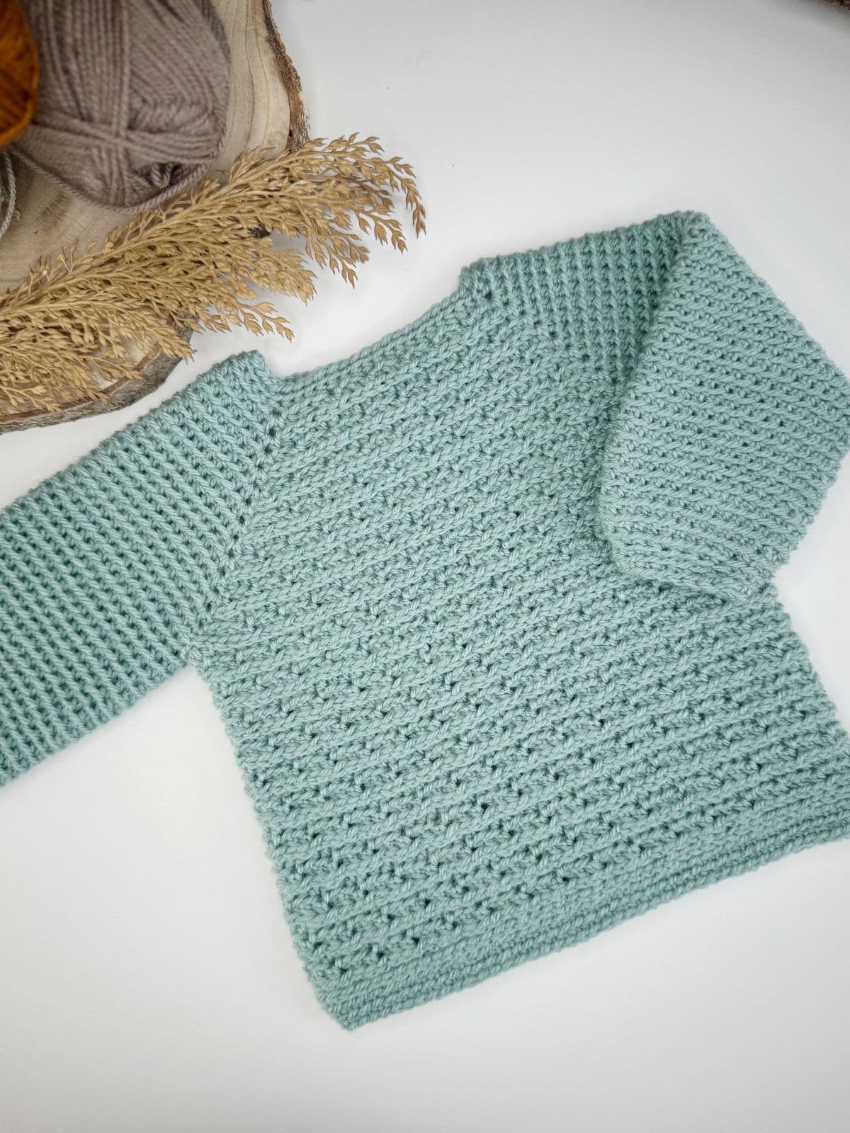 A handmade light blue crocheted sweater rests on a white surface, evoking the charm of creating an easy child crochet sweater. In the background, a basket brims with colorful yarn.