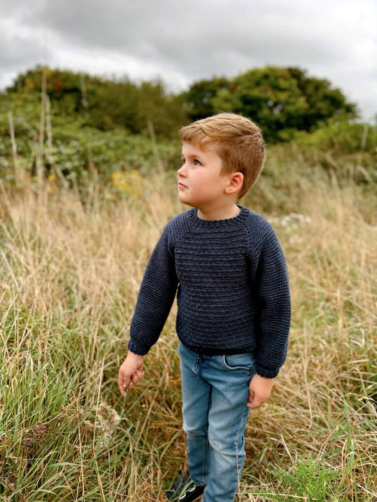 A young boy in a cozy, dark crochet sweater and jeans stands in a grassy field, looking to the side. Trees and a cloudy sky paint the backdrop.