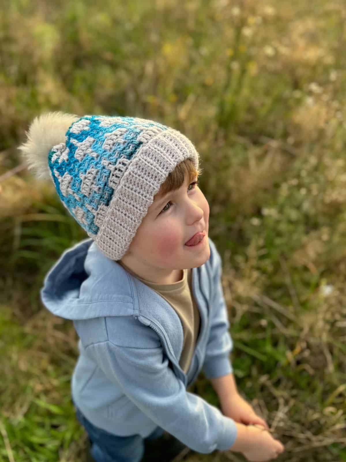 A child in a mosaic crochet hat with a pom-pom gazes upward, kneeling in a sunlit grassy field.