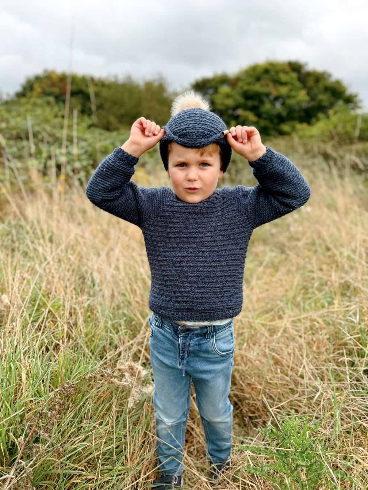 A child in an easy crochet sweater and jeans stands in a grassy field, adjusting a crochet hat.