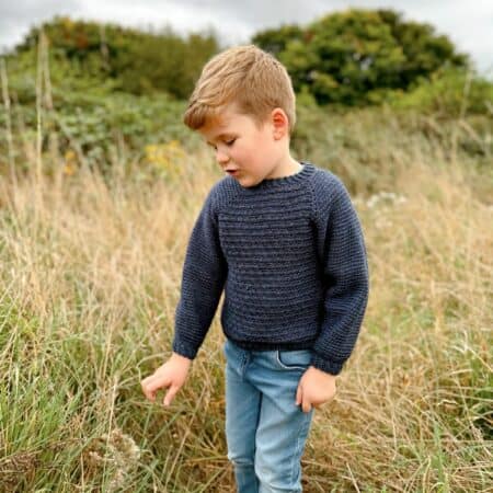 A young child in a navy crochet sweater and jeans stands in a grassy field, curiously looking down at plants. Tall trees loom in the background beneath a cloudy sky.