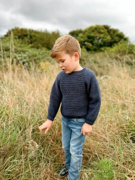 A young child in a navy crochet sweater and jeans stands in a grassy field, curiously looking down at plants. Tall trees loom in the background beneath a cloudy sky.