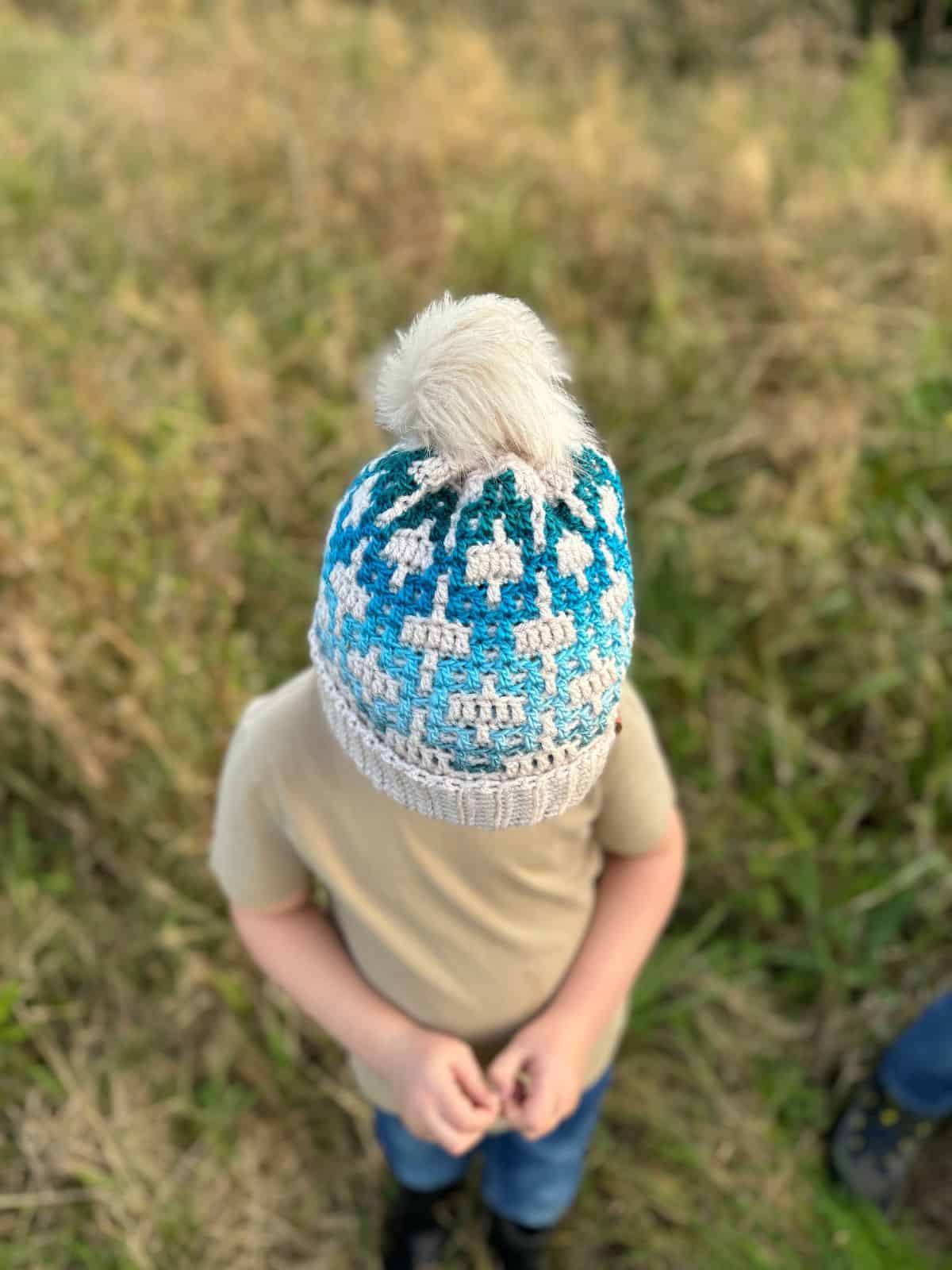 A child wearing a mosaic crochet hat, blue and white with a playful pom-pom, stands in a grassy field.