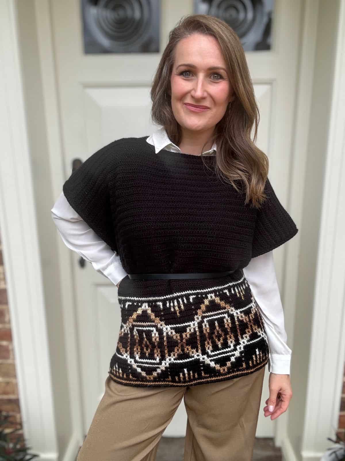 A woman stands in front of a door wearing a black crochet poncho over a white shirt, beige pants, and a patterned waist wrap that resembles an intricate crochet poncho pattern. She has long brown hair and a slight smile.