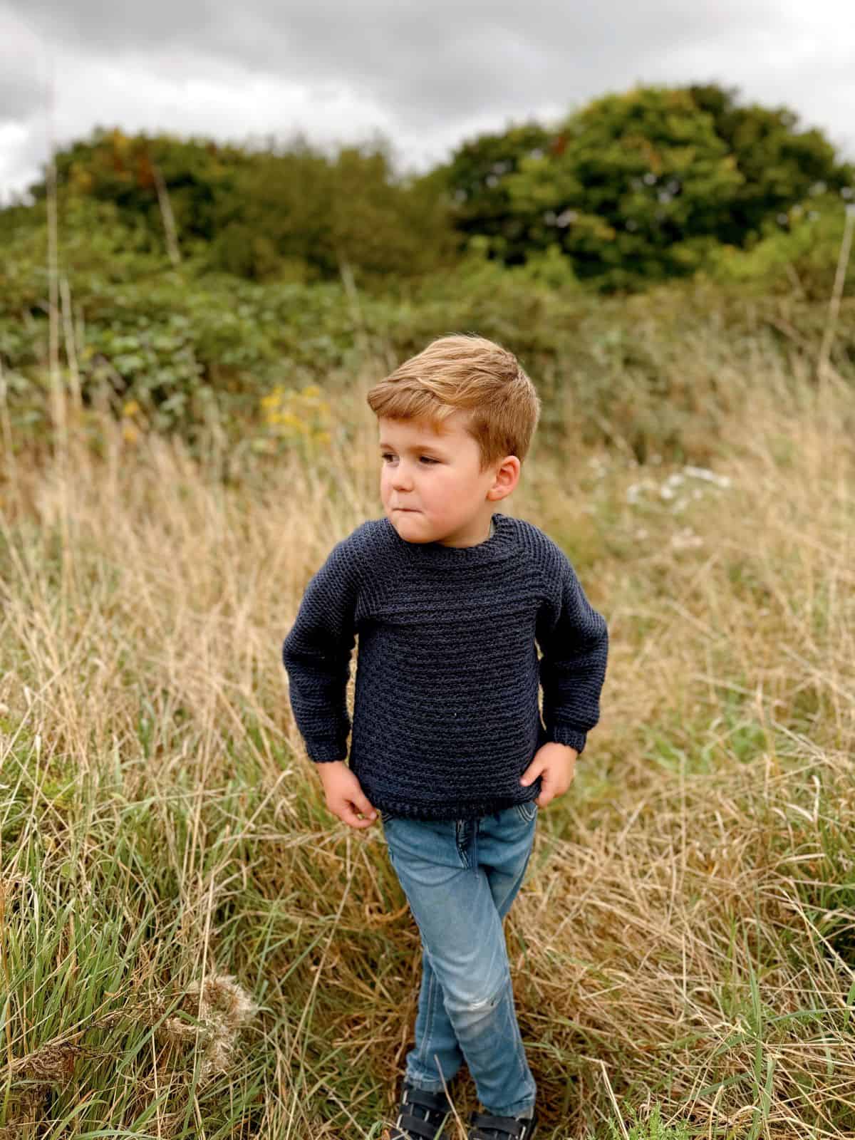 A young boy wearing a handmade navy crochet sweater, crafted with an easy child crochet pattern, stands in a grassy field with trees in the background.