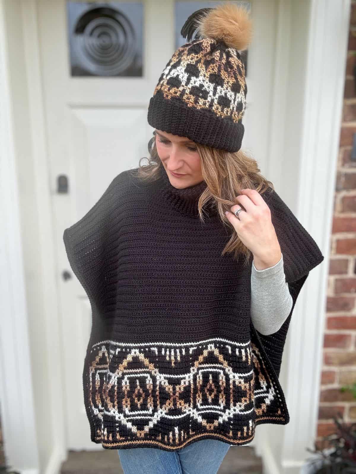 A person wearing a crochet poncho with crochet-inspired geometric patterns and a matching beanie stands in front of a white door and brick wall.