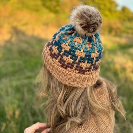 A person wearing a mosaic crochet hat with a pom-pom stands in a grassy field, facing away. The hat displays a geometric pattern in brown, blue, and beige.