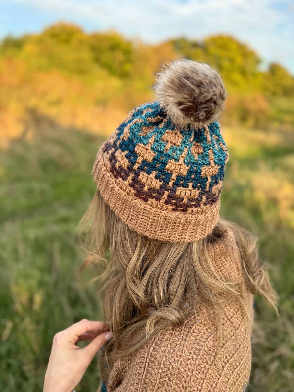 A person wearing a mosaic crochet hat with a pom-pom stands in a grassy field, facing away. The hat displays a geometric pattern in brown, blue, and beige.