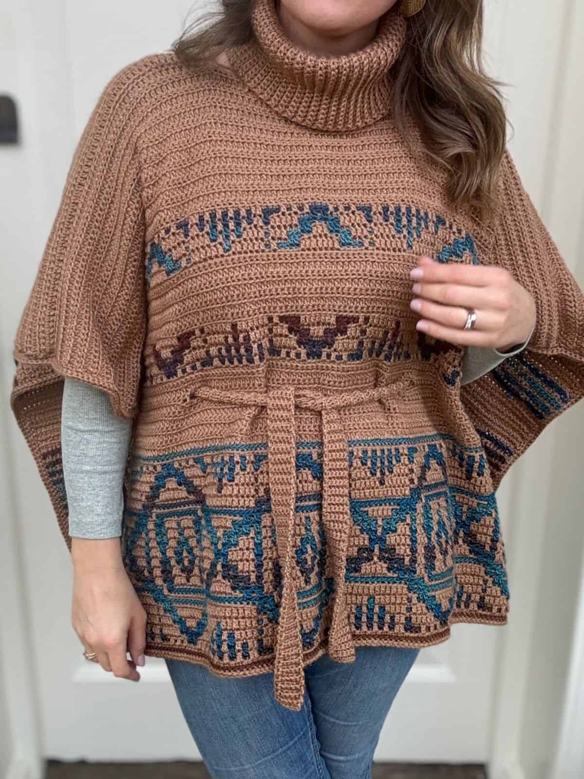 A person is wearing a stylish crochet turtleneck poncho, showcasing a crochet poncho pattern with geometric designs in brown and teal, paired effortlessly with a gray long-sleeve shirt and jeans.