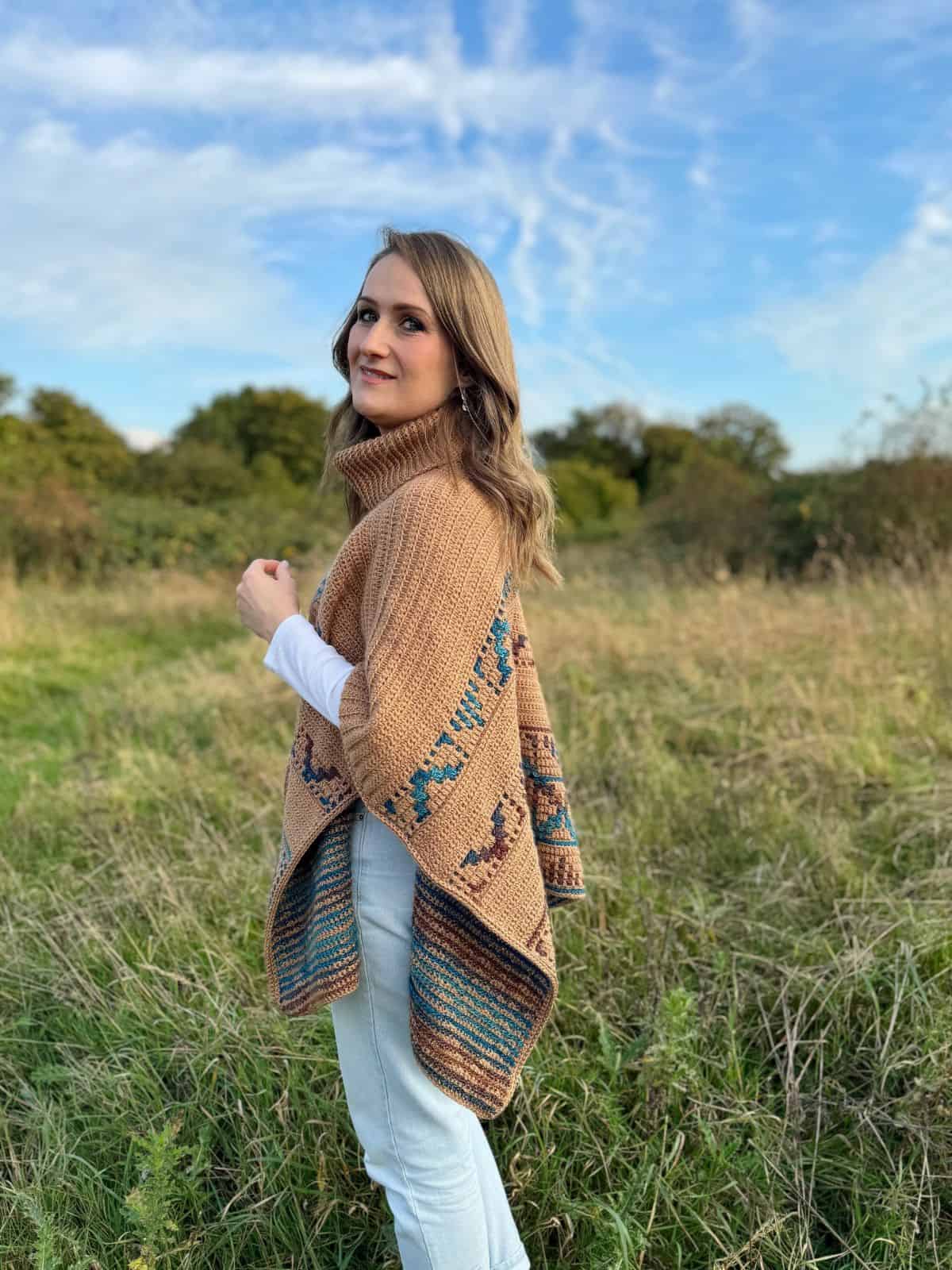 Woman standing in a field, draped in a brown crochet poncho with blue patterns reminiscent of a crochet poncho pattern, looks to the side under a blue sky with clouds.