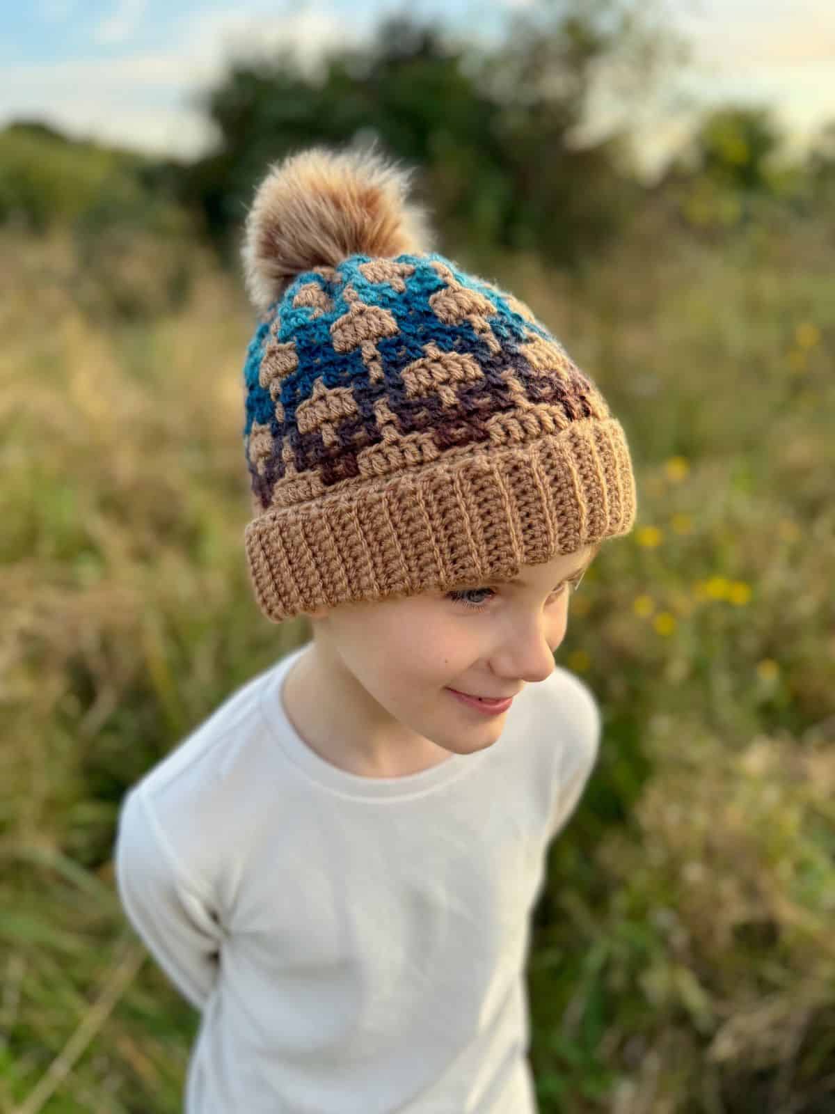 A child wearing a mosaic crochet hat with a pom-pom stands in a field, their expression neutral and thoughtful.