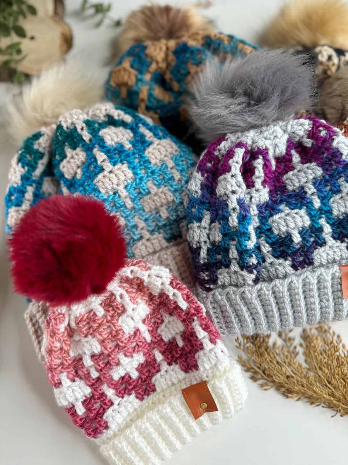 Colorful mosaic crochet beanies with pom-poms are arranged neatly, featuring blue, gray, pink, and cream tones.