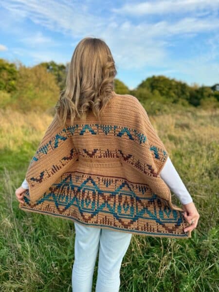 A person stands with their back turned, draped in a crochet poncho pattern of earthy browns, set against a backdrop of a grassy field, where trees meet the expansive blue sky.