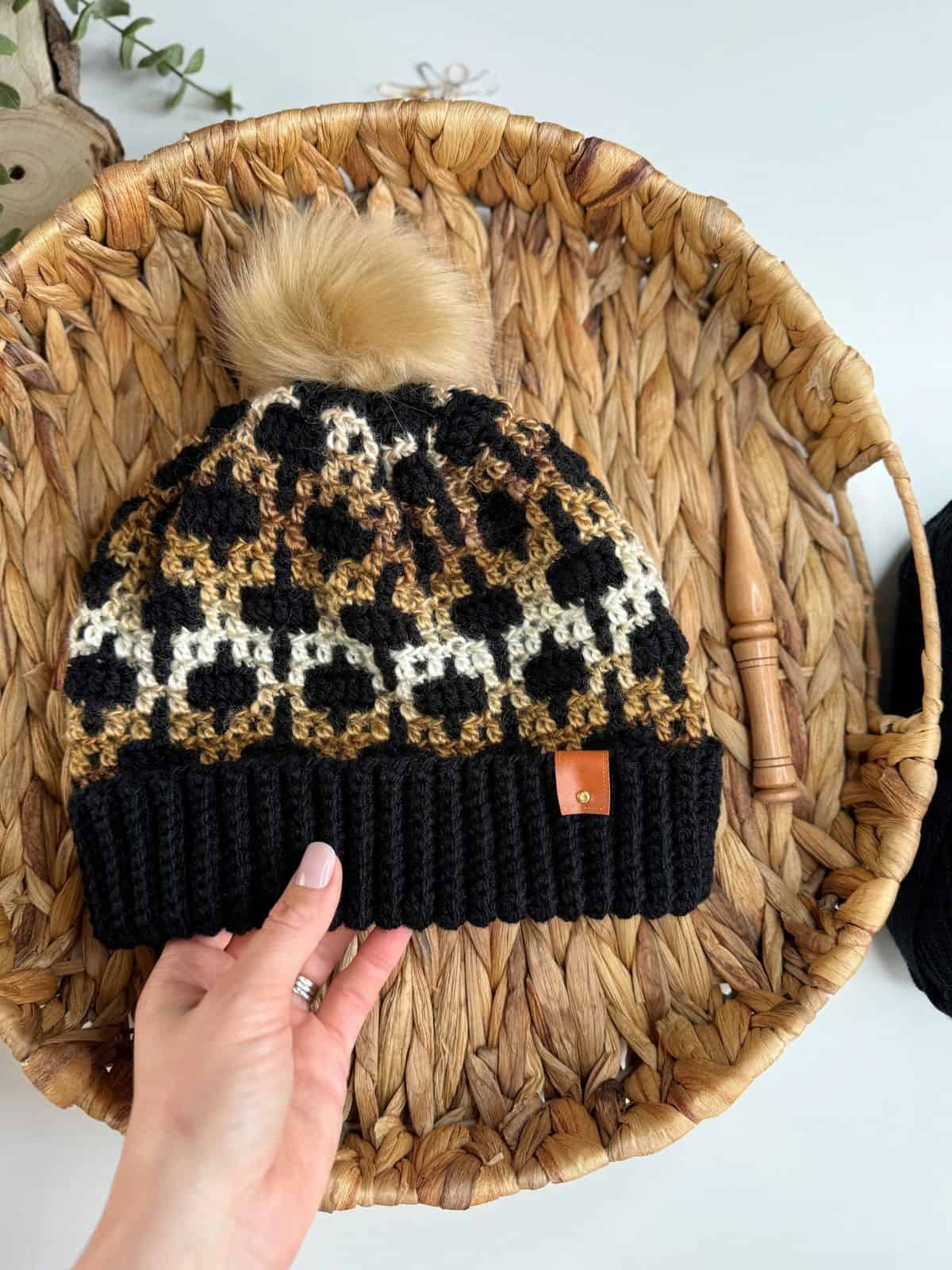 A hand holds a mosaic crochet hat adorned with a leopard pattern and pom-pom, displayed elegantly in a woven basket alongside crochet hooks and yarn.