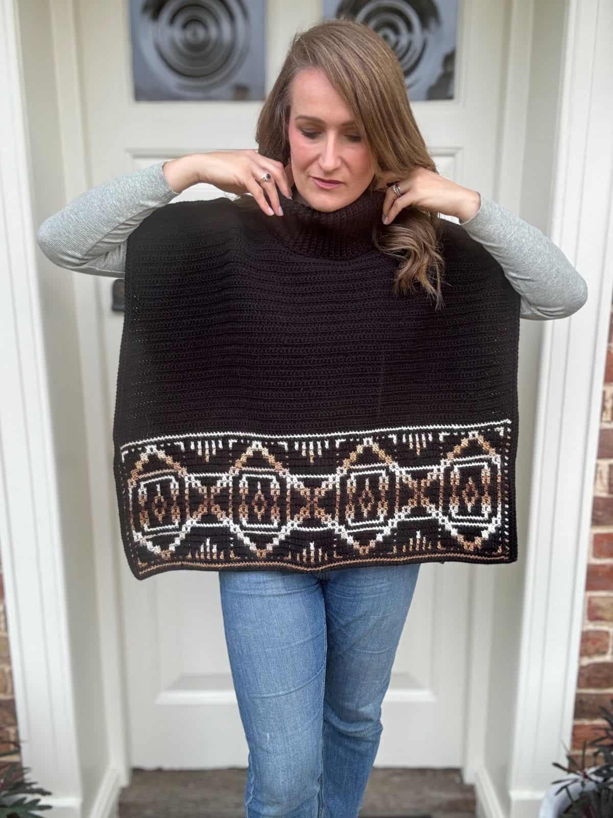 A person wearing a black crochet poncho with a geometric crochet pattern stands in front of a white door.