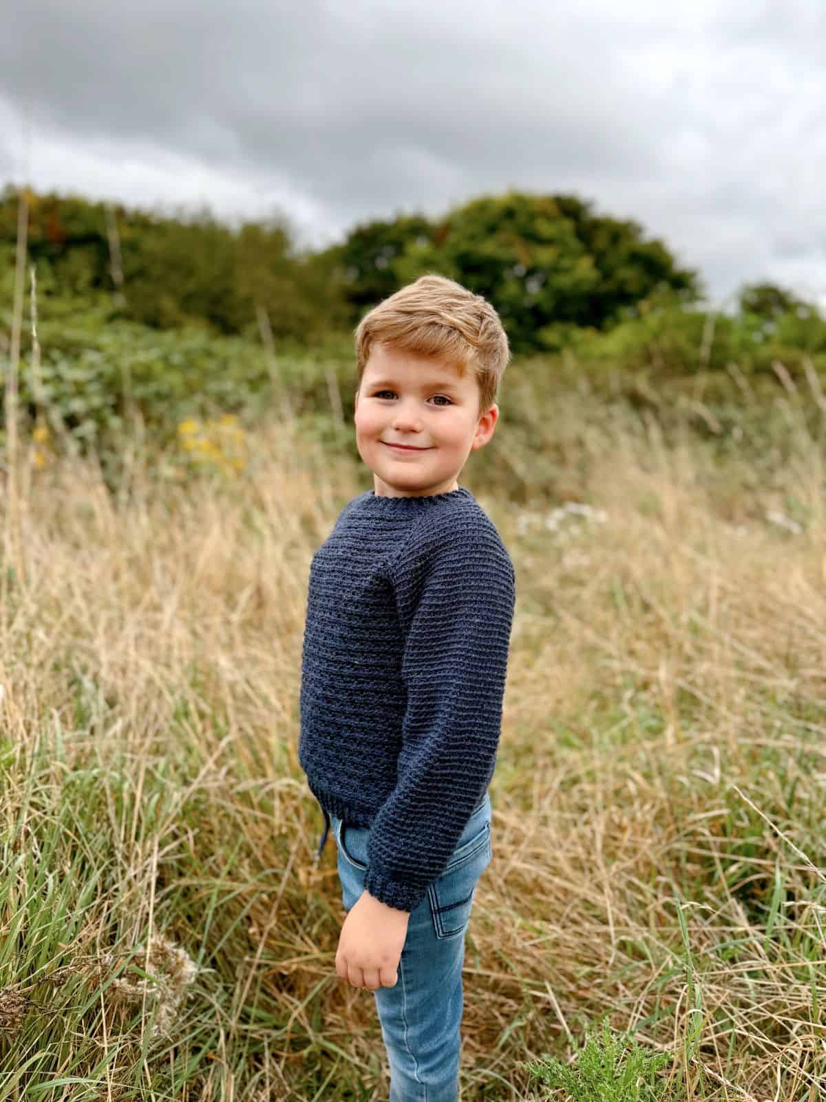 In a grassy field with greenery in the background, a child stands wearing jeans and an easy crochet sweater in blue.