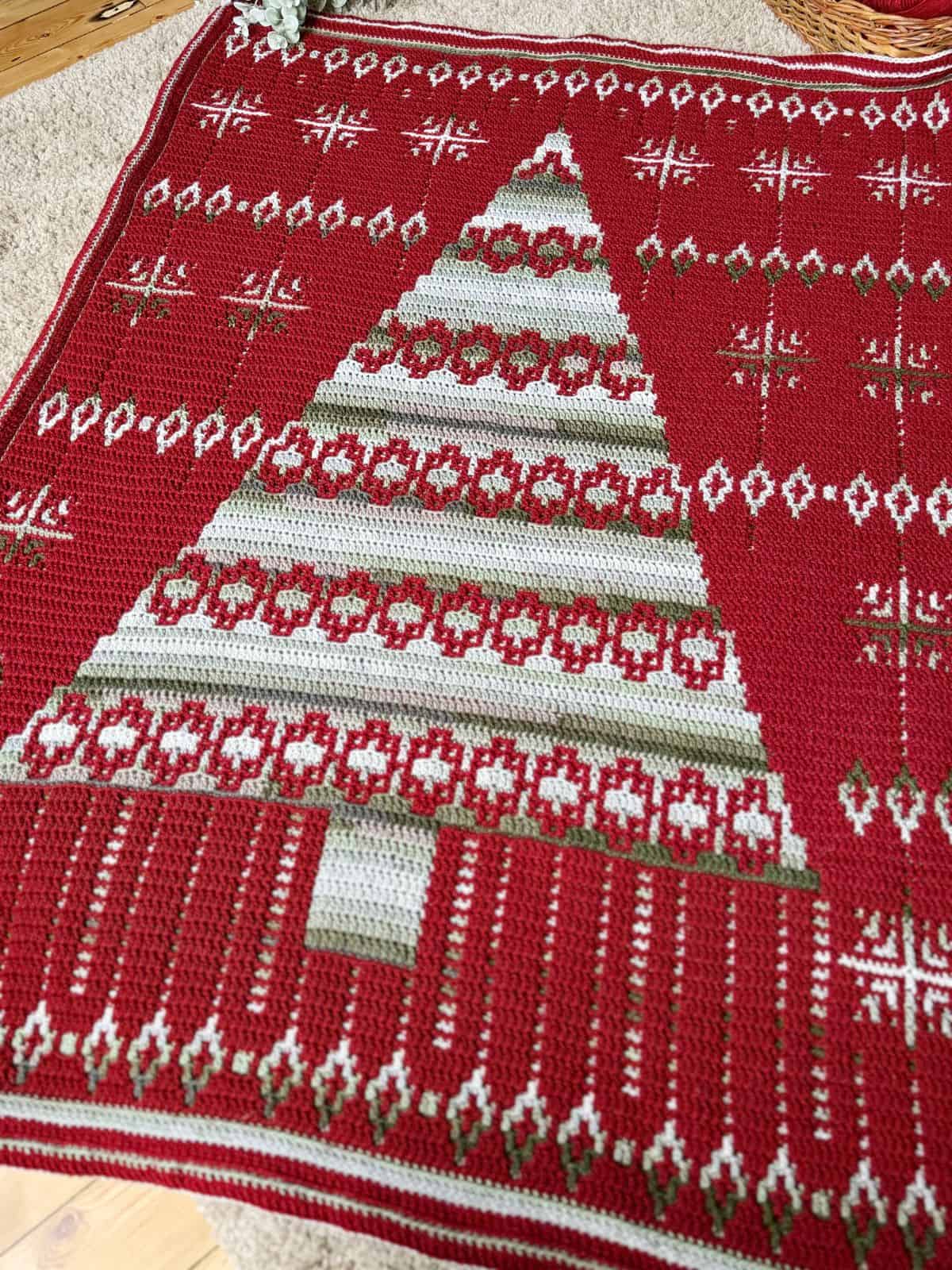 A red crochet Christmas blanket showcases a festive tree design adorned with white snowflakes and intricate geometric patterns, capturing the holiday spirit.