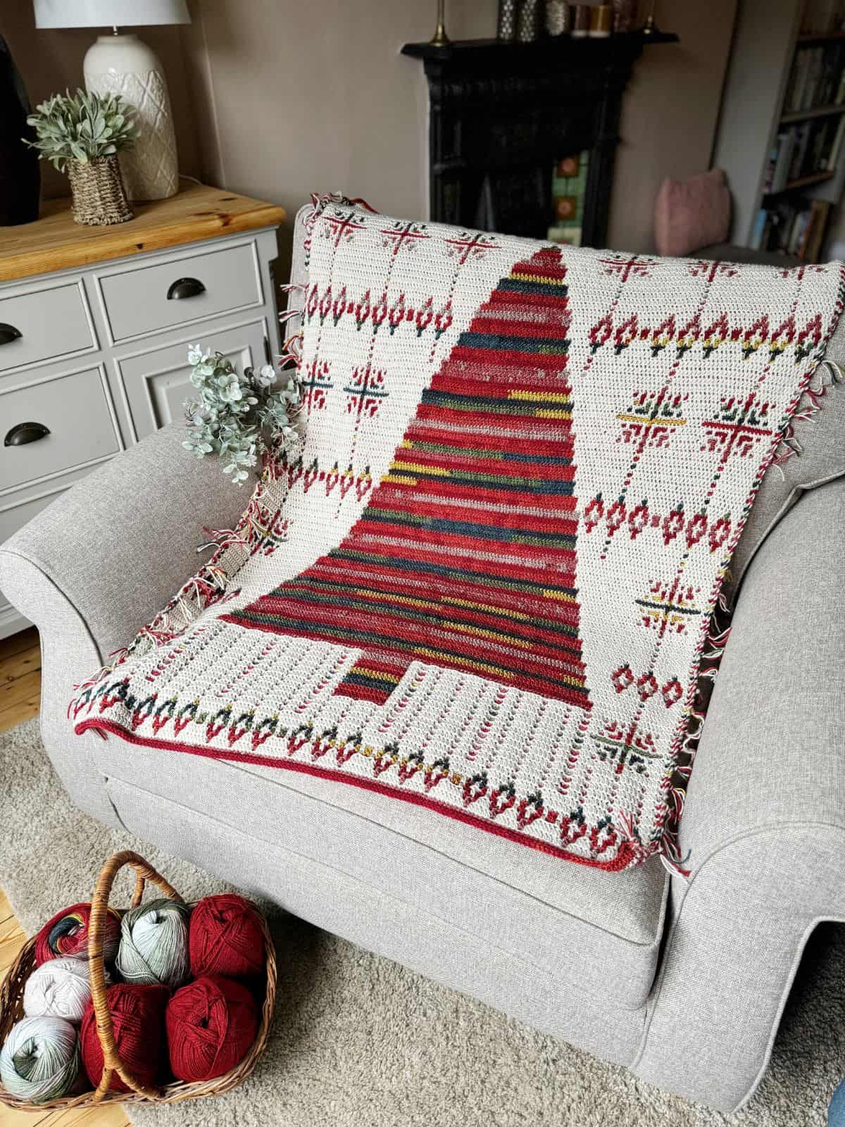 A cozy crochet Christmas blanket with a red and green tree pattern is draped over a gray armchair. Nearby, balls of yarn rest in a basket, ready for your next project.