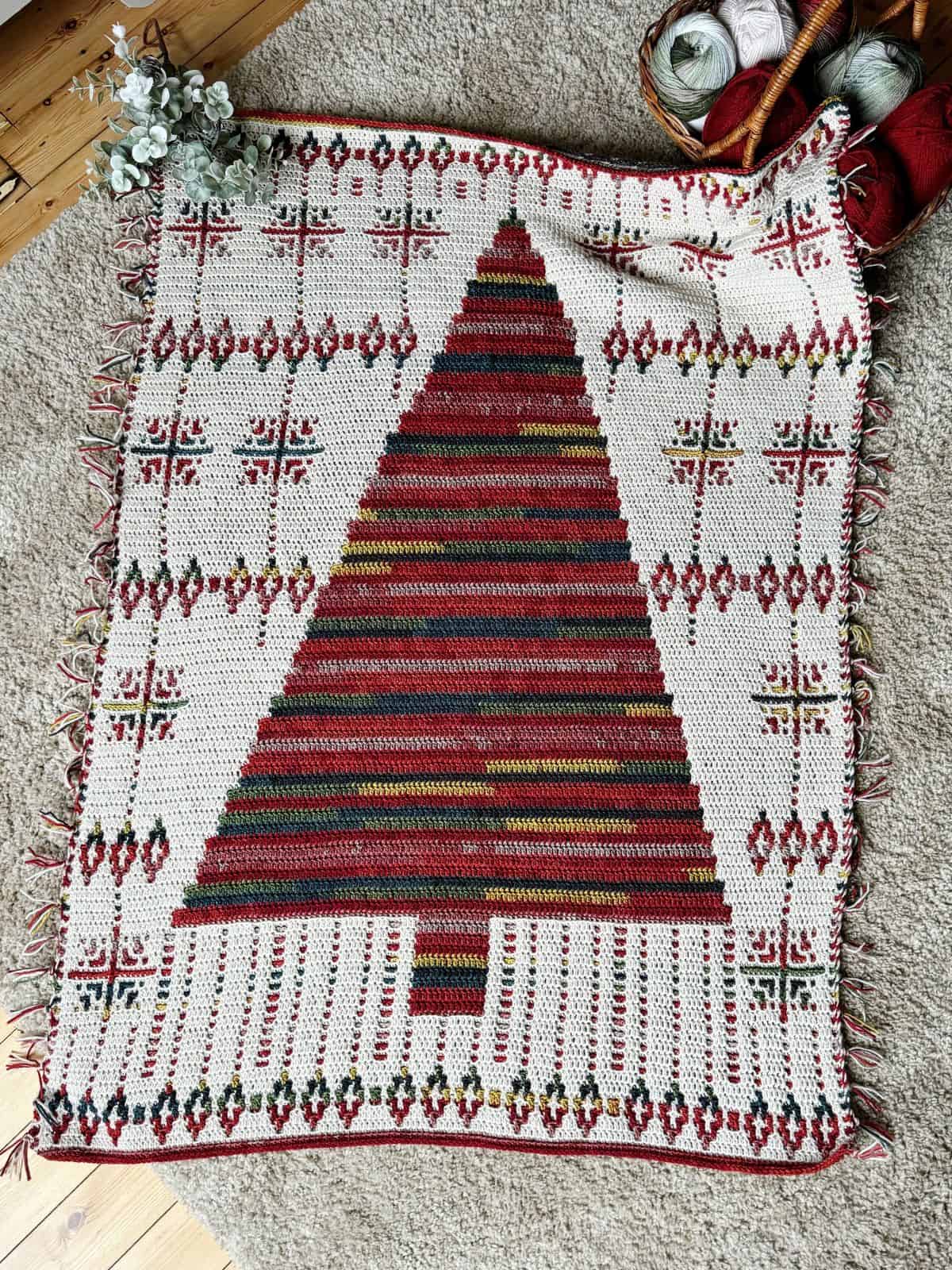 A crochet Christmas blanket featuring a central tree in red and brown stripes, surrounded by festive geometric designs. Crocheted edge details add a cozy touch.