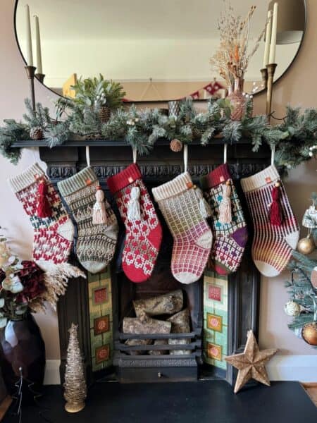 Five charming Christmas stockings, crafted with crochet Christmas stocking patterns, hang from a decorated fireplace mantel adorned with lush greenery and flickering candles.