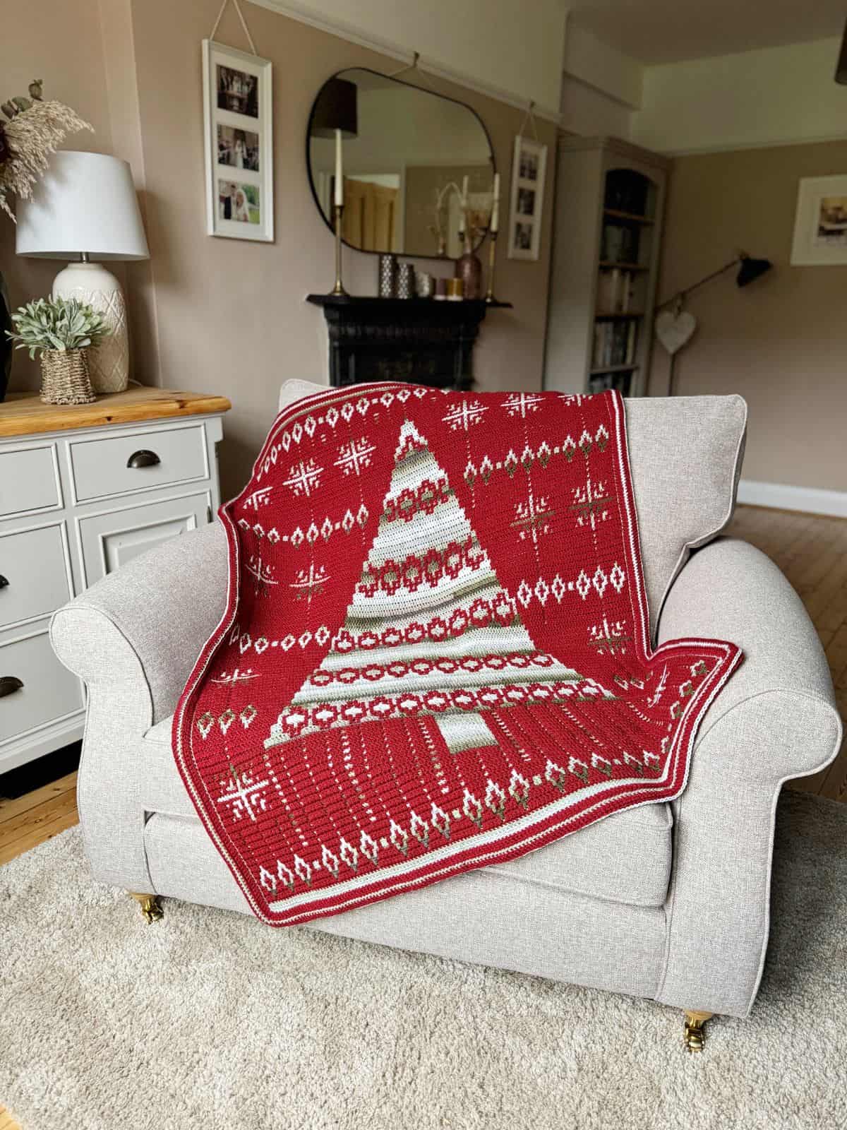 A red and white crochet blanket with a Christmas tree pattern draped over a beige armchair in a cozy living room.