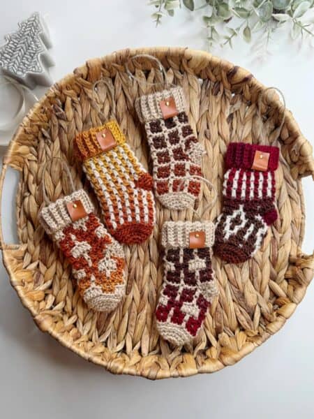 A woven basket holds five mini crochet Christmas stockings, each showcasing diverse patterns and colors. These charming stockings, accented with buttons, rest elegantly on a white surface.