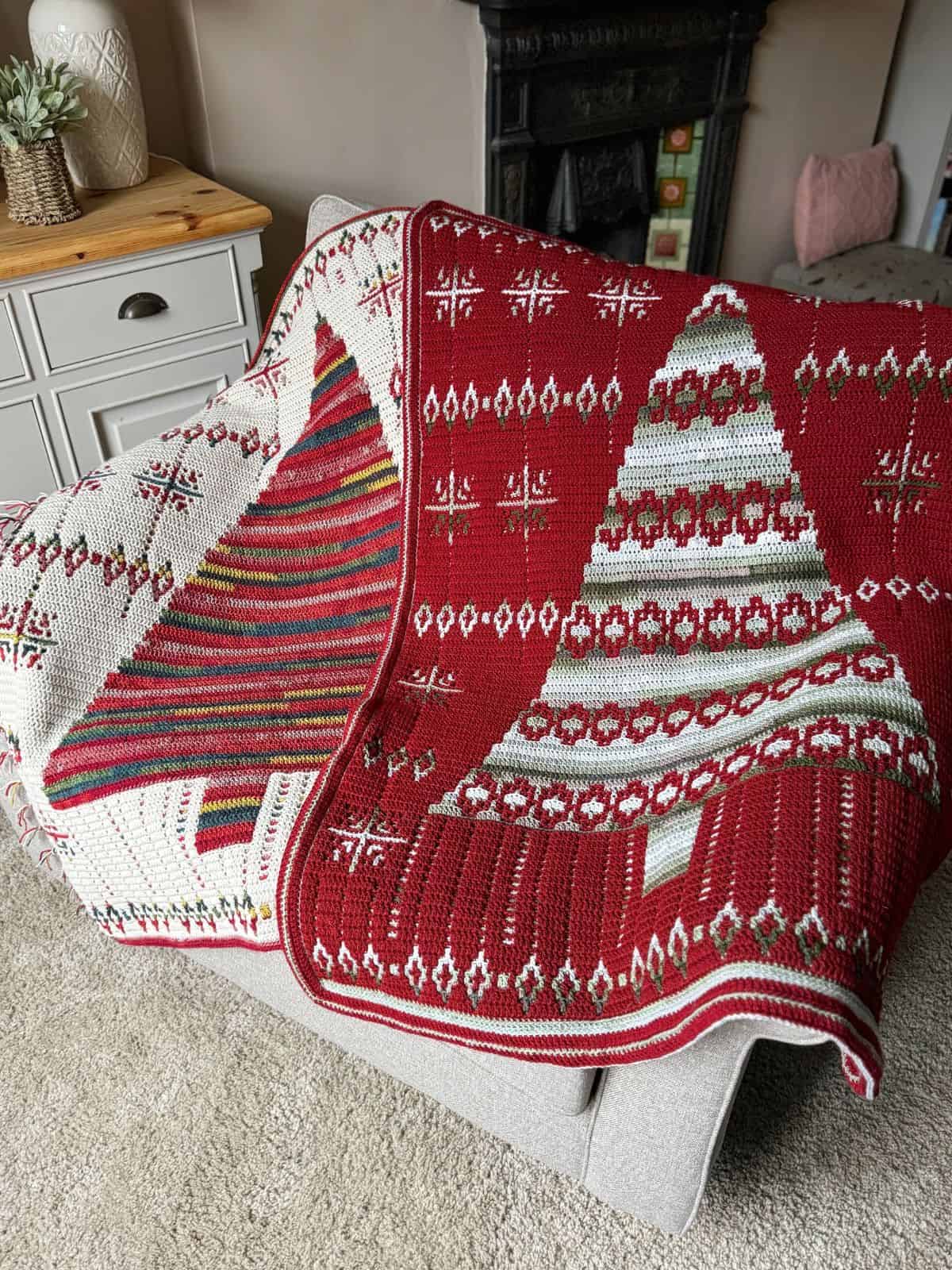 2 red and white crochet Christmas blankets pattern, adorned with festive tree designs, drapes elegantly over a gray chair in the cozy living room setting.