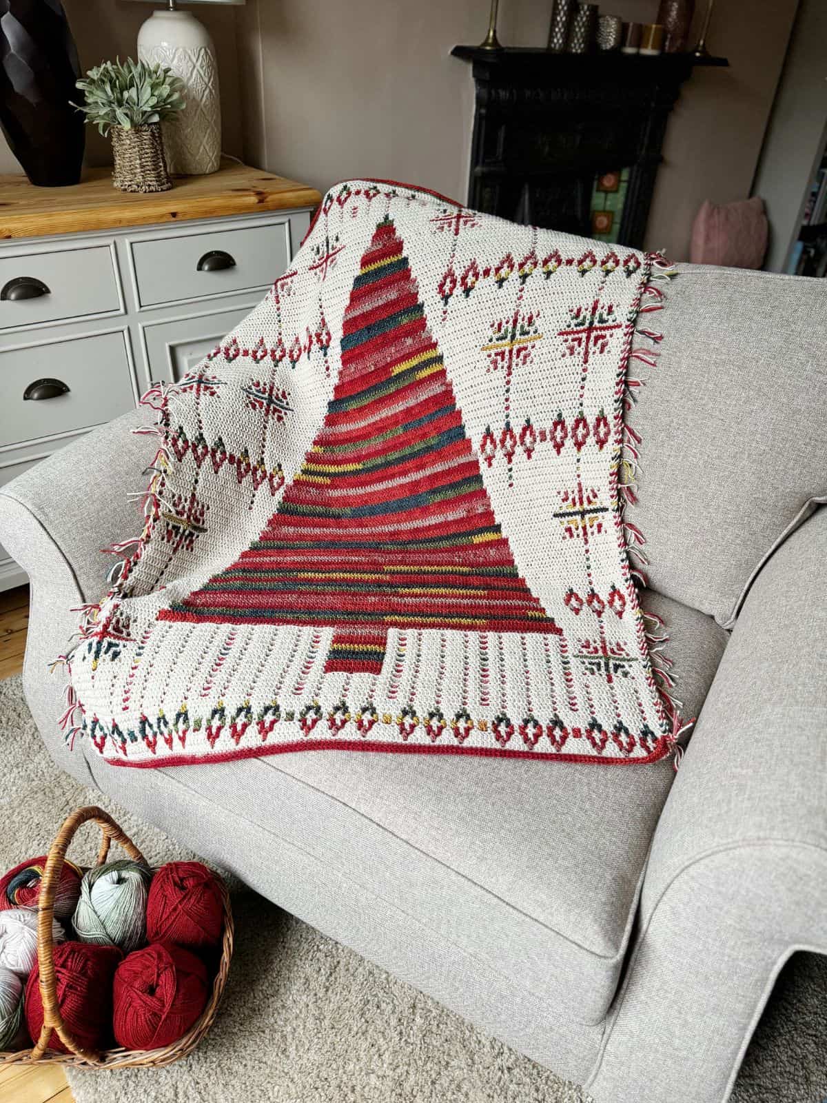 A beautifully crocheted Christmas tree blanket, featuring red, green, and white patterns, is draped over a gray armchair. Nearby, a basket with yarn balls hints at the creative process behind this festive crochet pattern.