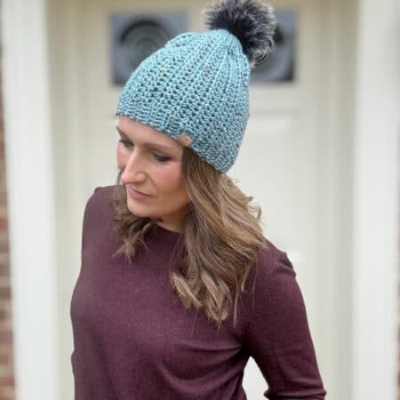 A person wearing a light blue crochet hat with a pom-pom and a maroon top, standing in front of a door.