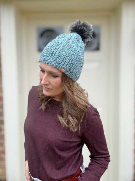 A person wearing a light blue crochet hat with a pom-pom and a maroon top, standing in front of a door.