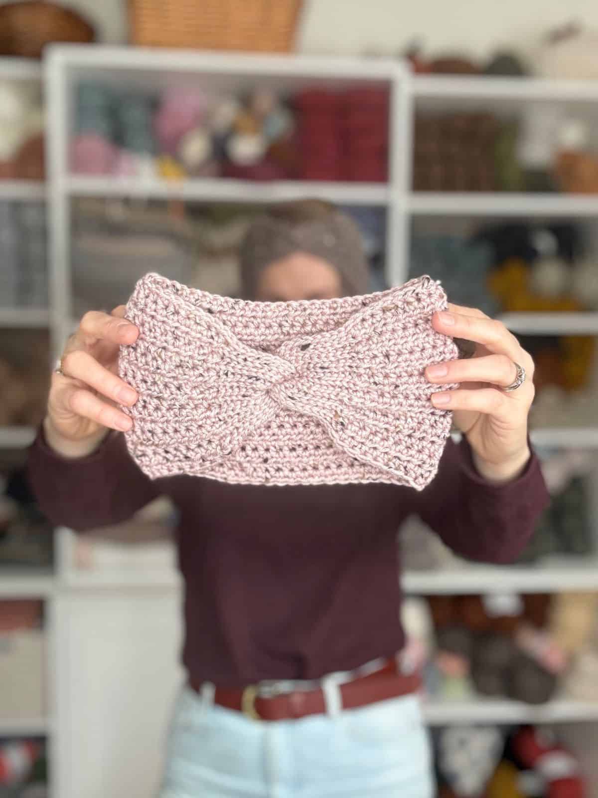 A person displays an easy crochet ear warmer pattern, showcasing a crocheted headband against a shelf brimming with vibrant yarn.