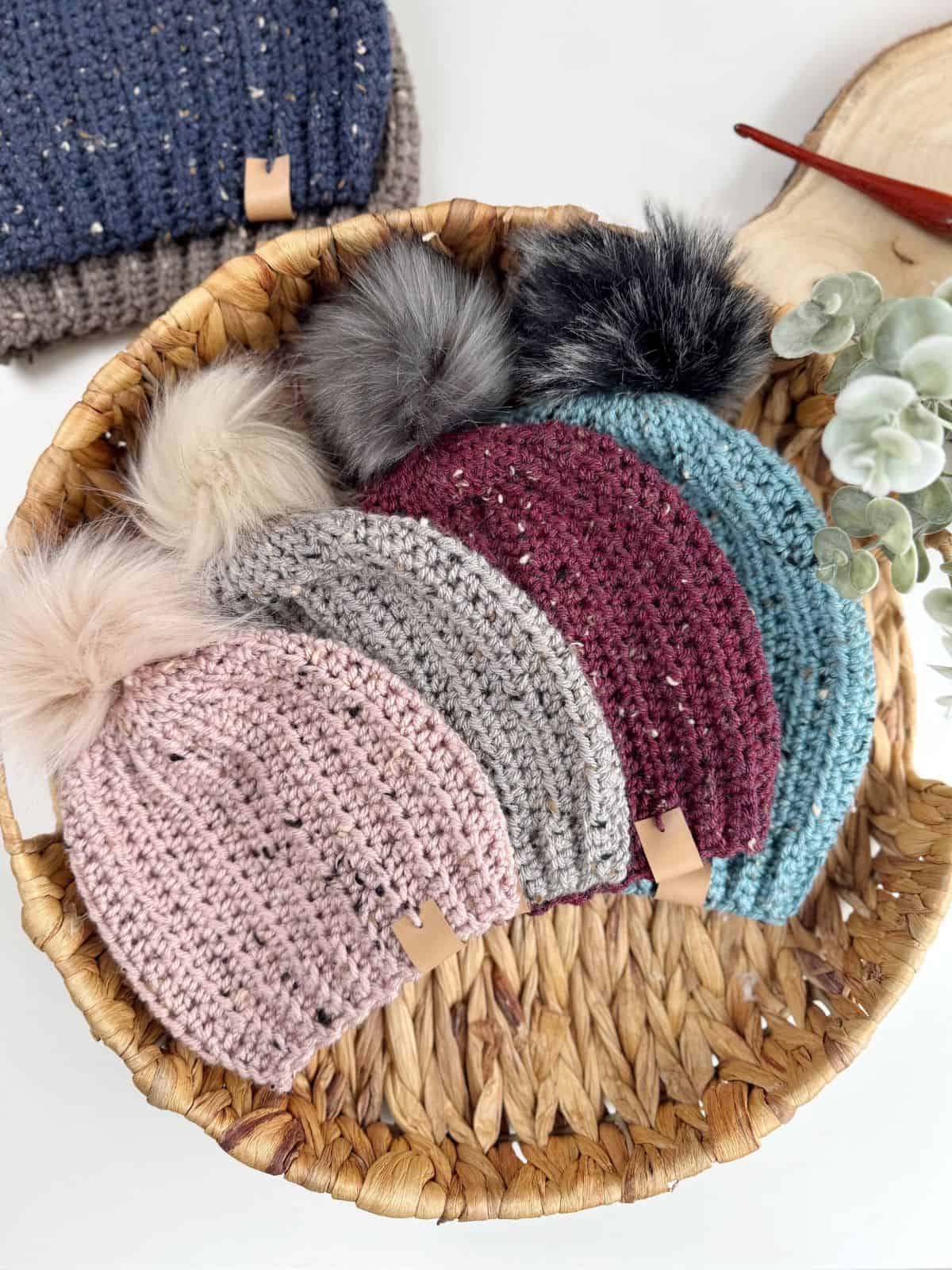 A basket holds five charming crocheted pom-pom beanies in pink, gray, maroon, teal, and dark blue—each inspired by an easy crochet hat pattern.