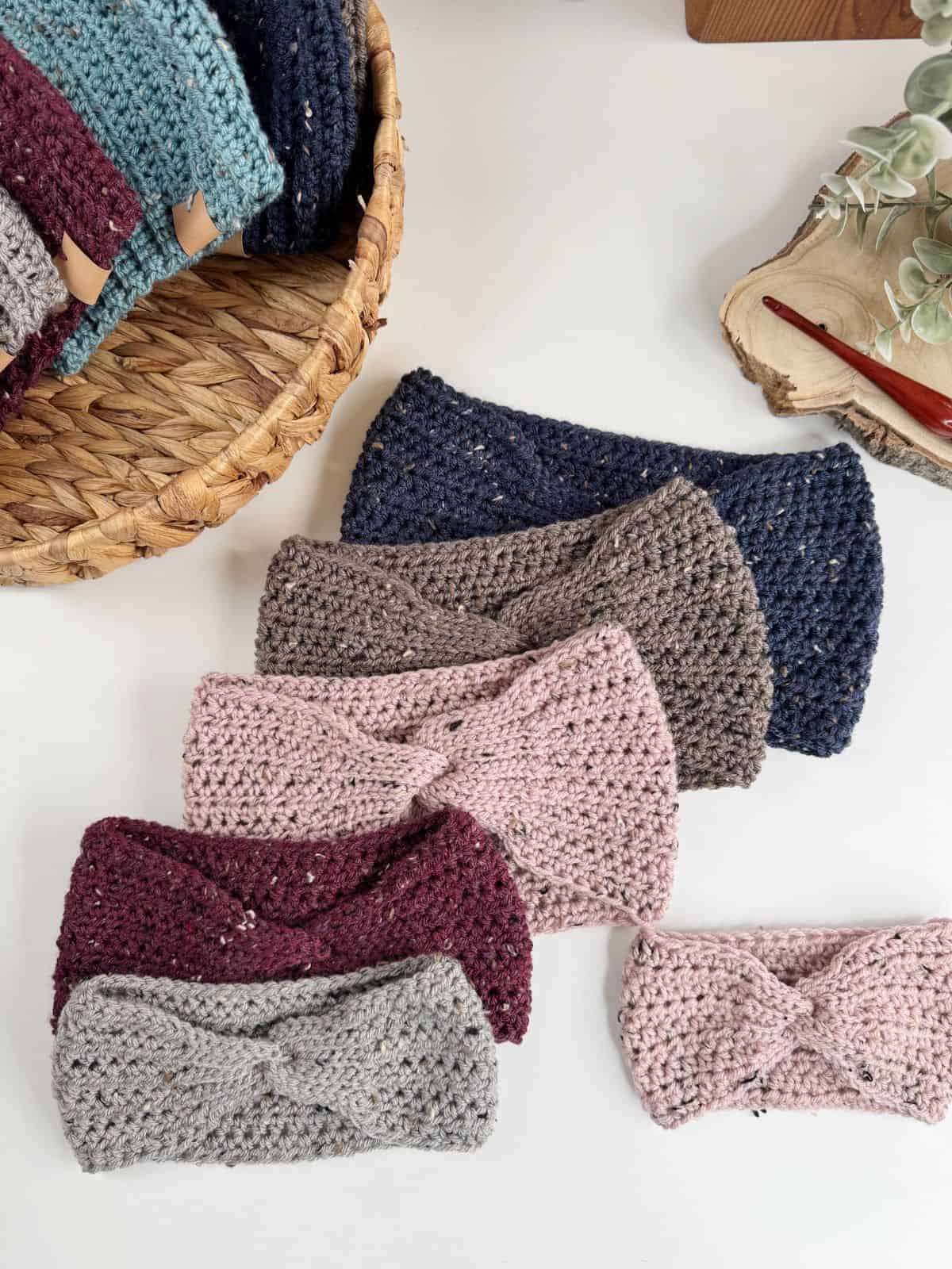 A display of crocheted headbands, crafted from an easy crochet ear warmer pattern, showcases a range of colors including pink, burgundy, gray, and blue. Set elegantly on a table beside a wicker basket and decorative foliage.