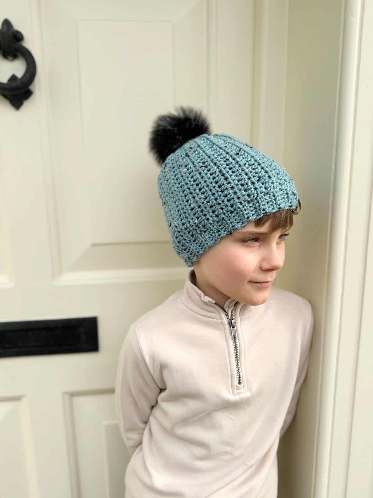 A child wearing a blue crochet hat with a black pom-pom, reminiscent of an easy crochet hat pattern, leans against a white door, looking to the side.