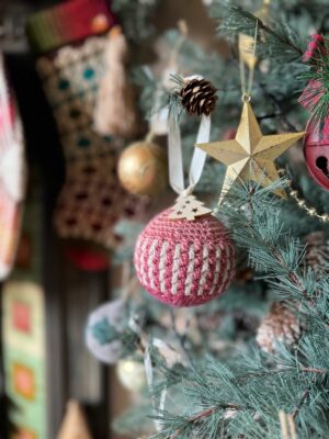 Close-up of a Christmas tree adorned with a pink and white crochet ornament, gold star, and pinecone. Blurred in the background, crochet tree decorations hang alongside stockings and other festive embellishments.