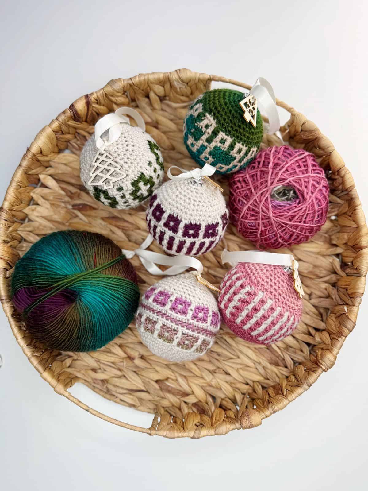 A wicker basket holds six crocheted Christmas tree decorations and two balls of yarn in green and pink hues.