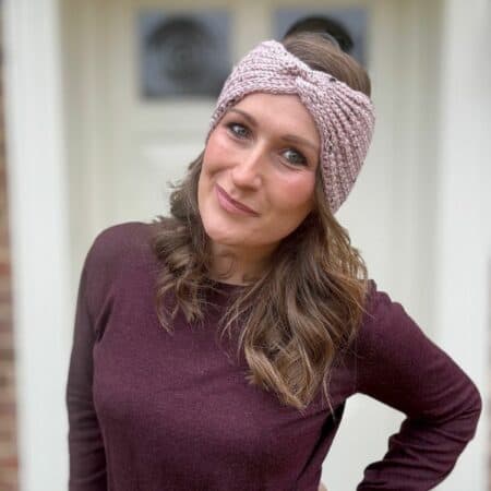 A woman with long hair, wearing a purple sweater and an easy crochet ear warmer pattern as a textured pink headband, stands outside, smiling with her hand on her hip.