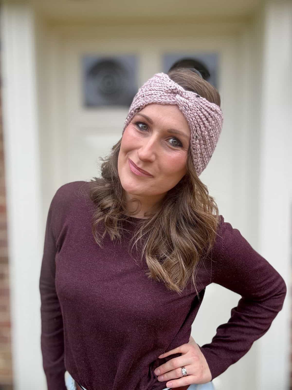 A woman with long hair, wearing a purple sweater and an easy crochet ear warmer pattern as a textured pink headband, stands outside, smiling with her hand on her hip.