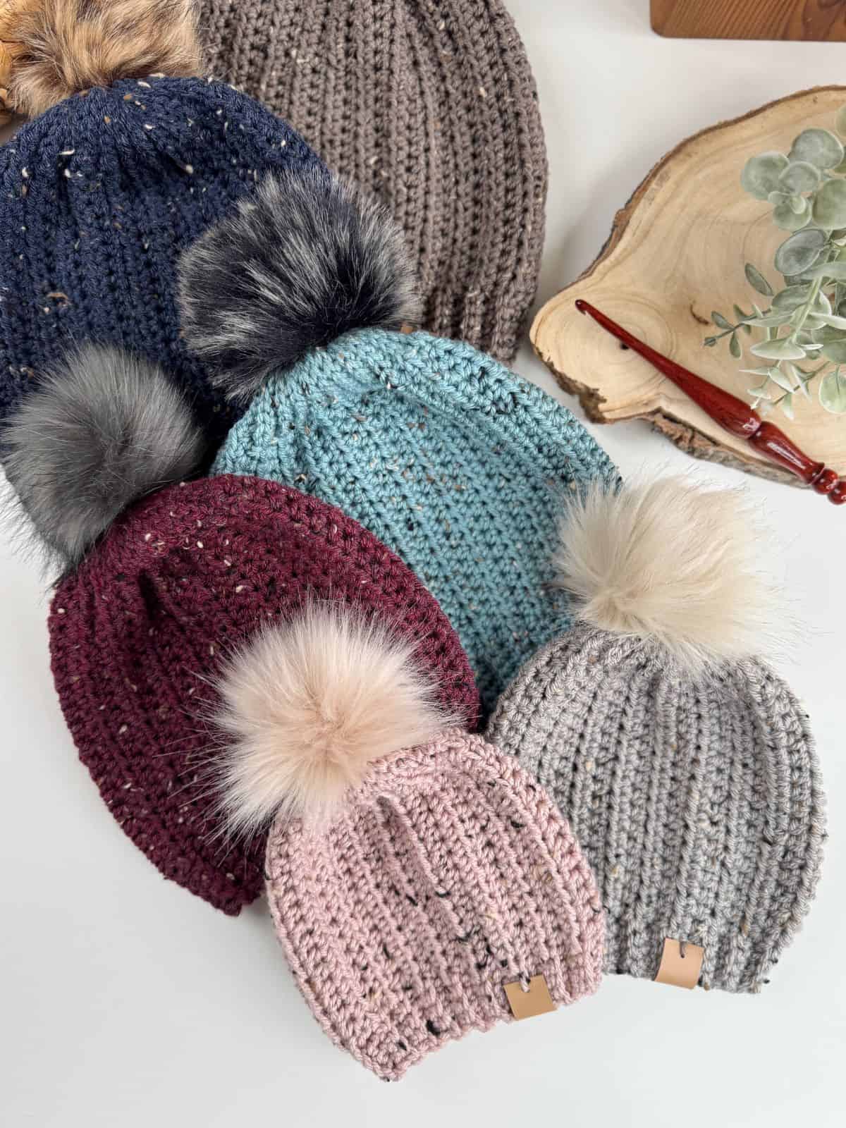 A collection of crochet hats with pom-poms in various colors, reminiscent of an easy crochet hat pattern, is displayed on a white surface alongside a wooden tray with eucalyptus and a red pen.