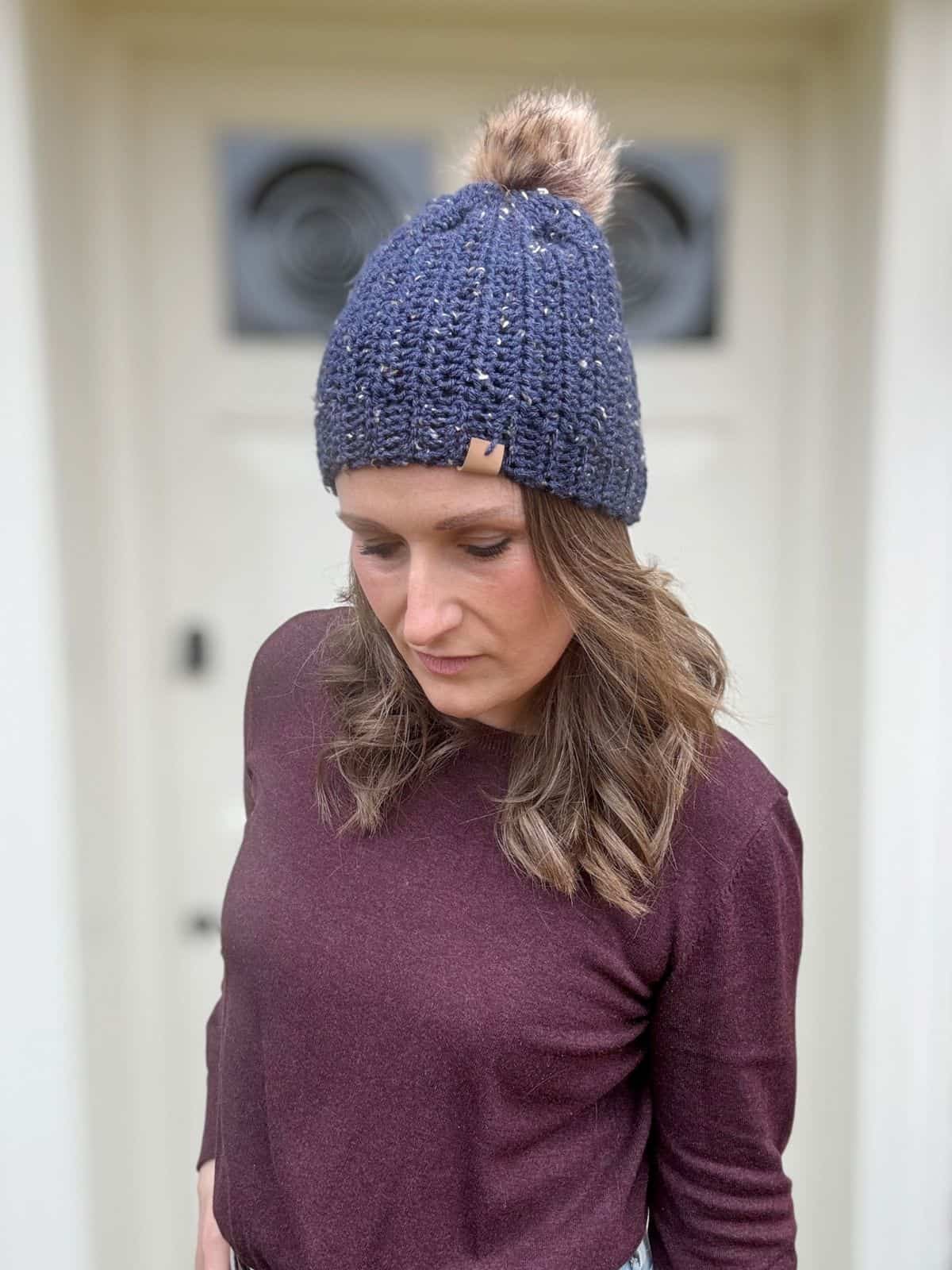 A woman in a blue crochet beanie with a pom-pom, crafted from an easy crochet hat pattern, gazes downward while standing in front of a light-colored door.