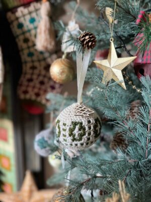 A Christmas tree adorned with crochet tree decorations features charming ornaments, including a crocheted ball with the number "24," a golden star, pinecones, and other festive elements.