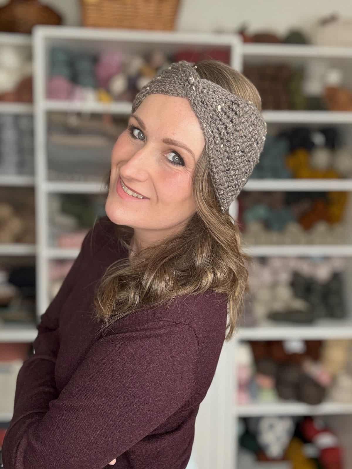 A person wearing a crocheted headband and a dark long-sleeve top smiles with arms crossed. Yarn and craft supplies, reminiscent of an easy crochet ear warmer pattern, are visible in the background.