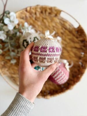 A hand holding a crocheted ornament with a geometric pattern, nestled among crochet tree decorations on a woven tray.